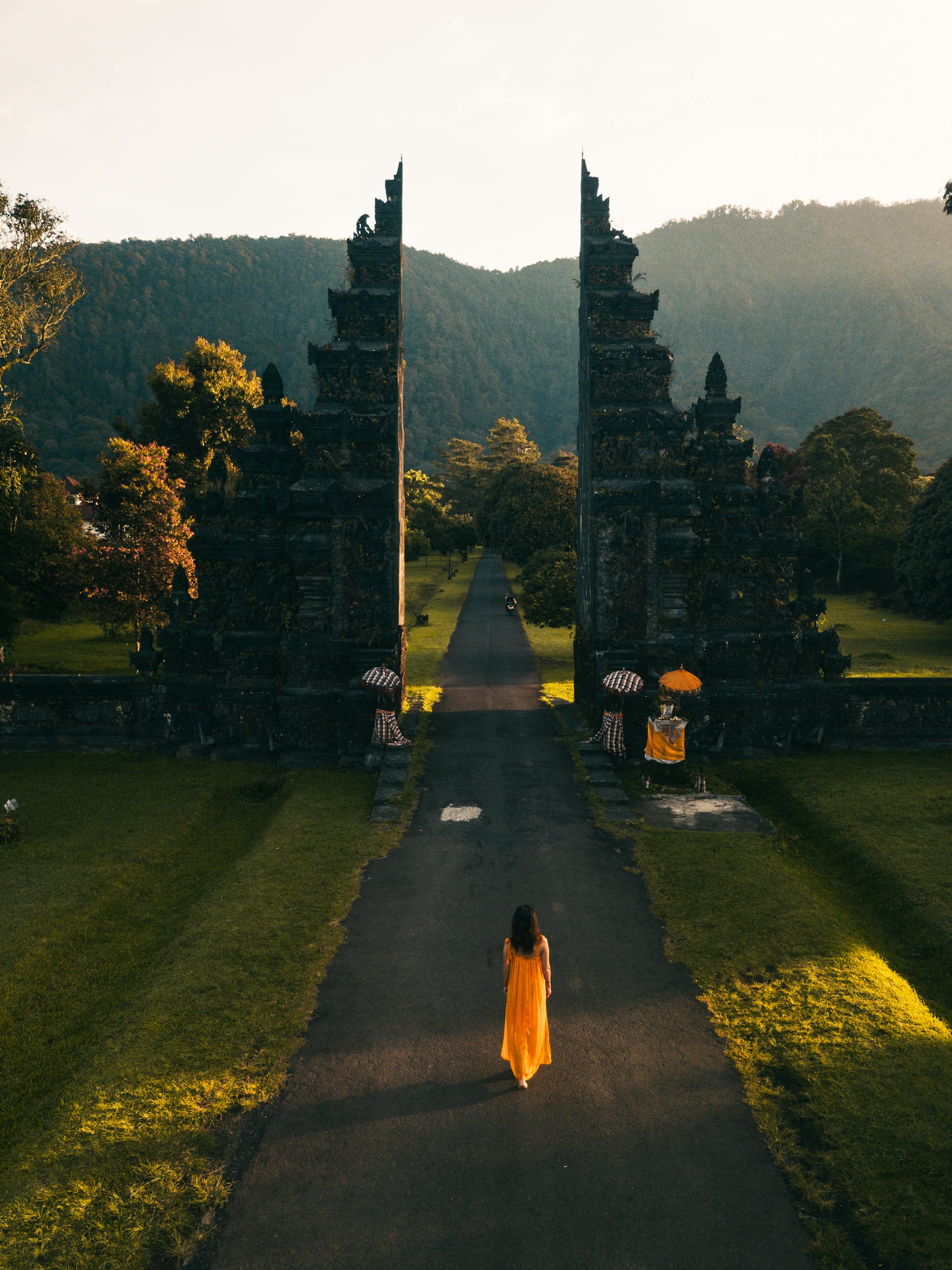 Aerial view of a woman in orange dress walking towards Bali Handara Gate at sunrise in Indonesia.